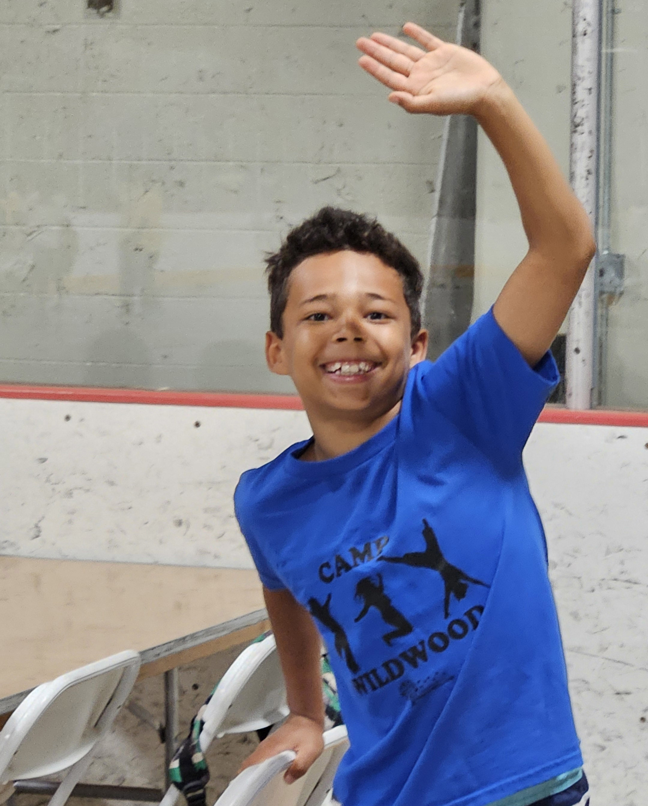 A young male Camp Wildwood camper in a blue camp shirt smiles and waves at the camera