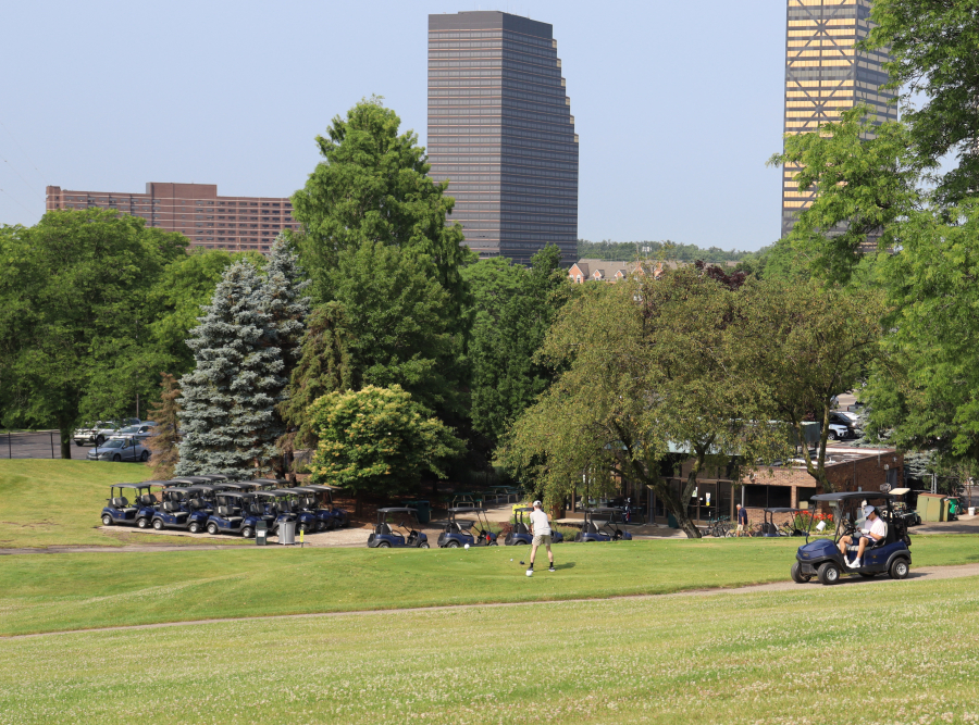 Golfer teeing off at Evergreen Hills Golf Course with a golf cart nearby, clubhouse and Southfield skyline visible in the background.