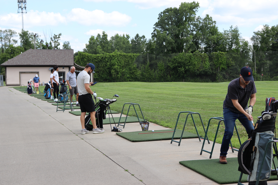 Golfers practice their swings from mat tee stations at Beech Woods Driving Range. Several individuals stand along the driving range hitting area with golf bags nearby, taking practice shots toward the open grassy fairway. Trees and the driving range building appear in the background.